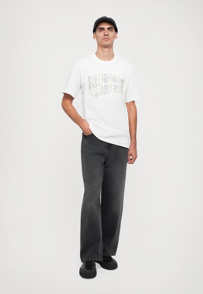 Tricou alb cu mâneci scurte, având un grafic "Billionaire Boys Club", asortat cu blugi largi gri închis și teniși negri, purtat cu o șapcă neagră.