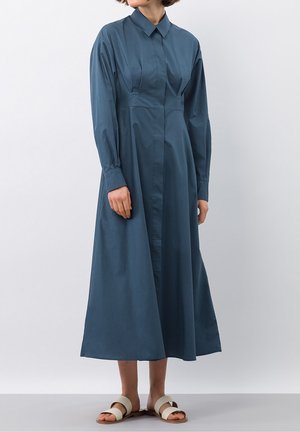 Donna che indossa un vestito midi blu scuro a maniche lunghe, con bottoni e colletto, e sandali beige bassi, in piedi davanti a uno sfondo semplice.