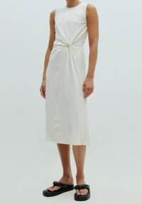 Robe midi blanche sans manches avec un détail torsadé à l'avant, portée avec des sandales à plateforme noires, modèle debout sur un fond uni.