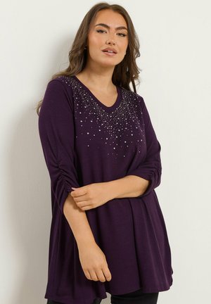 Tunique violette à manches longues avec un décolleté en V, ornée de détails argentés autour du col et de manches froncées. Tissu doux avec une coupe décontractée.