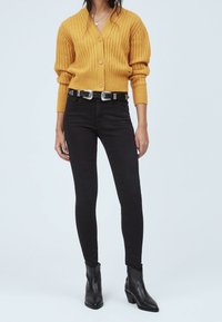 Kvinna iklädd en senapsgul ribbad kofta, svarta högmidjade skinny jeans med dubbel spänne och svarta ankelboots, stående.