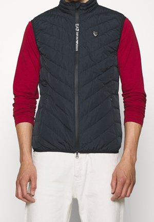 Mężczyzna ubrany w czarną pikowaną kamizelkę bez rękawów z logo EA7 Emporio Armani, na czerwonej koszuli z długim rękawem i białych spodniach.