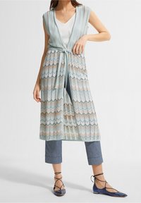 Cardigan aperto blu chiaro con cintura in vita, caratterizzato da un motivo a zig zag in beige, blu e bianco. Indossato sopra una maglietta bianca e jeans.