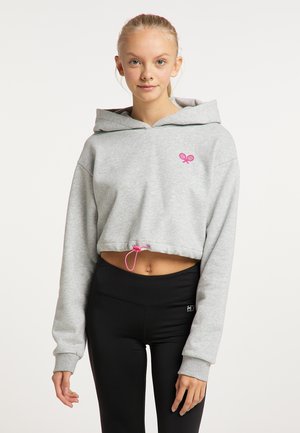 Jeune femme portant un sweat à capuche court gris avec un logo raquette de tennis rose et un legging de sport noir, debout devant un fond clair uni.
