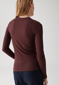Donna con capelli biondi che indossa un top a maniche lunghe attillato a coste di colore bordeaux e jeans blu scuro, vista di spalle su uno sfondo neutro.