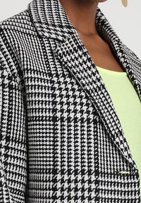 Blazer a motivo houndstooth bianco e nero con colletto a rever e design strutturato, abbinato a un top verde chiaro sottostante.