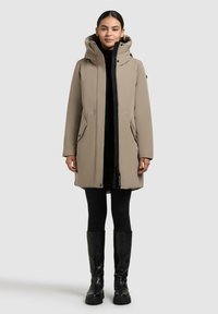Beige waterdichte jas met een capuchon, rits aan de voorkant, zijzakken en een strak ontwerp, gedragen over een zwarte coltrui en zwarte leggings.