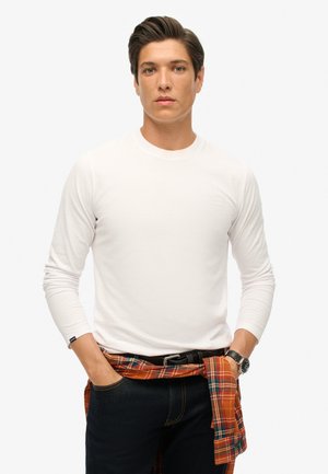 Witte langslijp T-shirt met ronde hals, zachte textuur, gecombineerd met donkere jeans en een geruite shirt om de taille geknoopt. Zwarte horloge zichtbaar.