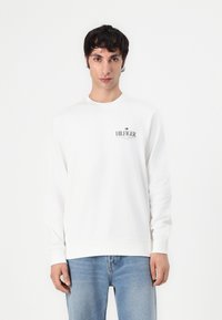 Hvit genser med rund hals, ribbestrikkede mansjetter og liv, med en trykt logo på venstre bryst. Båret med lyseblå jeans.