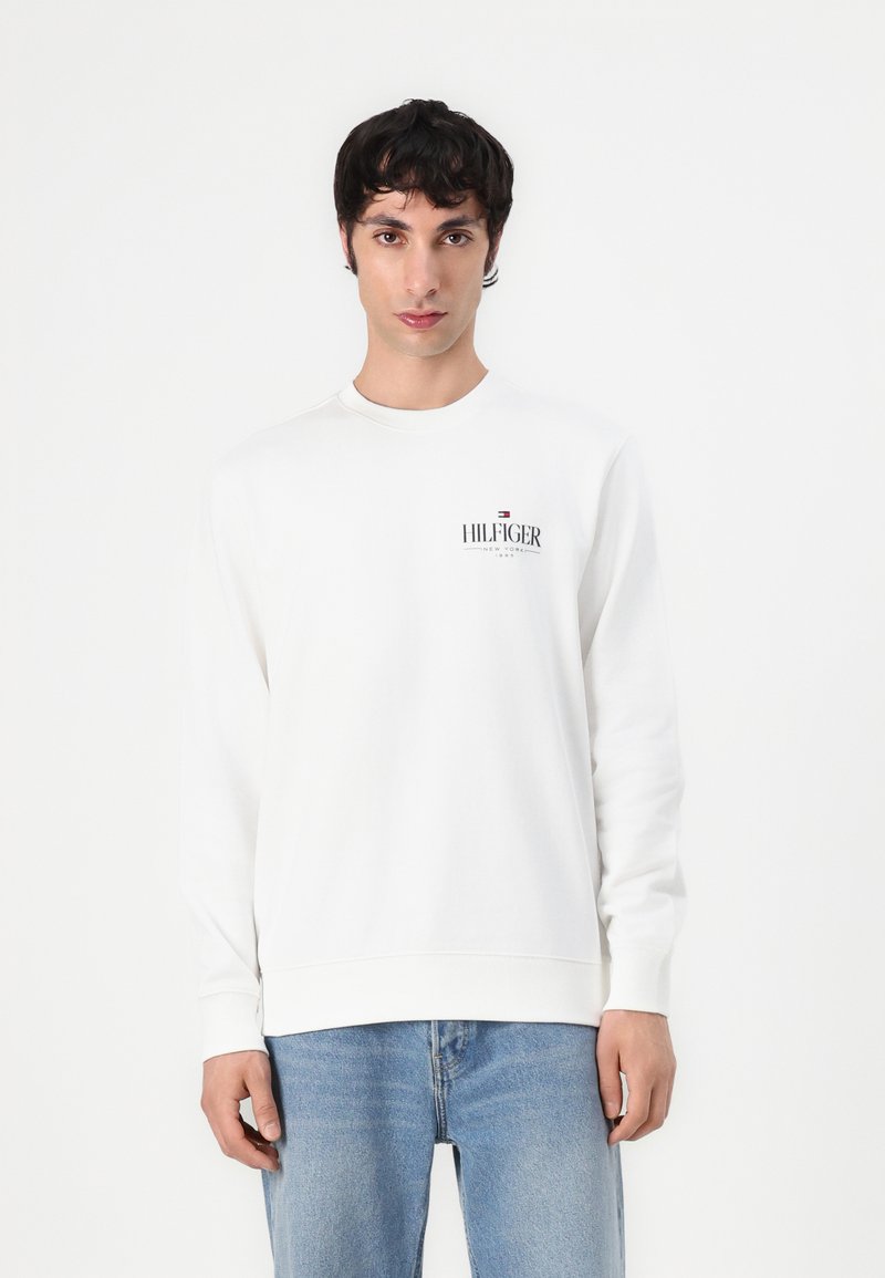 Hvit genser med rund hals, ribbestrikkede mansjetter og liv, med en trykt logo på venstre bryst. Båret med lyseblå jeans.
