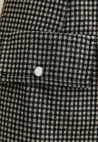 Il tessuto a scacchi bianco e nero presenta una superficie testurizzata, con una tasca a soffietto chiusa da un pulsante a scatto argentato.