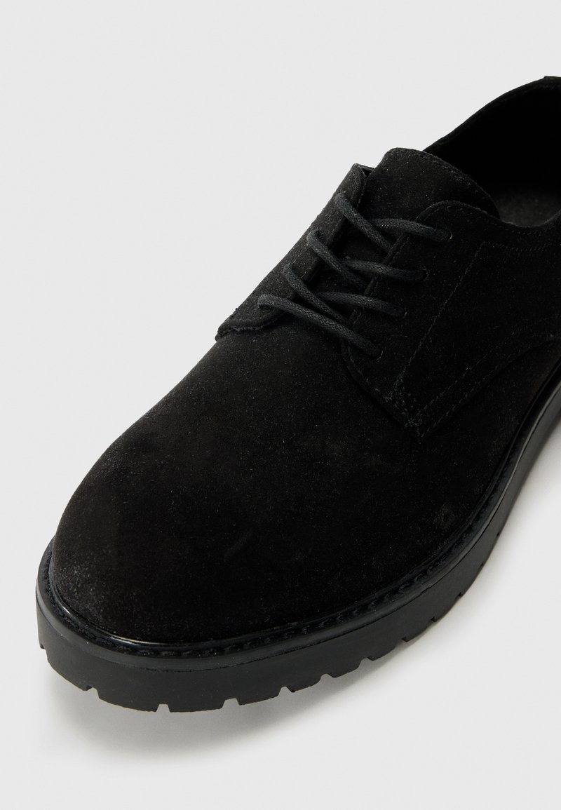 Chaussure en daim noir à lacets avec un bout arrondi, une texture lisse, une épaisse semelle en caoutchouc noir, et des détails de couture subtils le long de la tige.