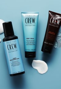 Drie American Crew verzorgingsproducten voor heren op een blauw oppervlak: verzorgingsschuim, vezelcrème en stylinggel met sterke fixatie, met productmonsters ernaast.