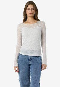 Jauna sieviete ar gariem brūniem matiem, tērpta caurspīdīgā baltā garajā piedurknē, kas uzlikta virs baltas kamizoles un zila džinsās, stāvot pret baltu fona.