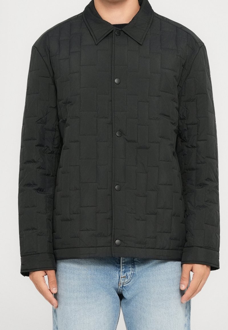 Homme portant une veste matelassée noire avec des boutons-pression et un jean bleu clair, debout devant un fond uni.