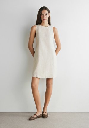 Frau steht vor einer weißen Wand, trägt ein ärmelloses beiges knielanges Kleid und braune gemusterte Ballerinas mit Riemchen.