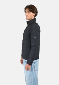 Doudoune noire avec des coutures horizontales, col montant, fermeture zippée et détail de logo sur la manche ; portée avec un jean en denim bleu clair.