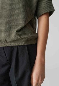 Top verde oliva dalla texture caratteristica con maniche corte, vestibilità comoda e orlo arricciato, abbinato a pantaloni neri larghi.