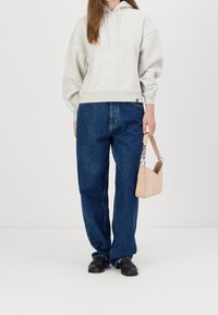 Šedá mikina s volným střihem a kapucí se šňůrkami, doplněná volnými modrými džínami. V jedné ruce drží malou béžovou kabelku.