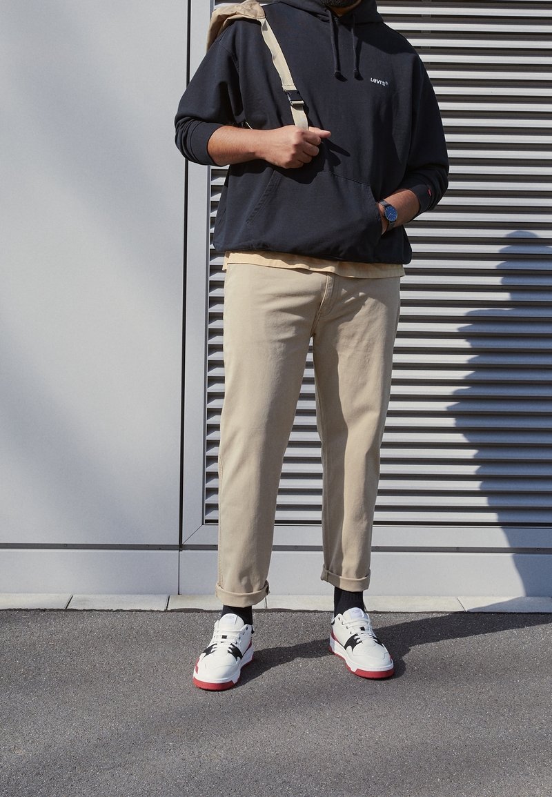 Personne debout à l'extérieur portant un pantalon beige, un hoodie noir avec logo, des baskets blanches à semelles rouges et un sac à dos beige sur une épaule.