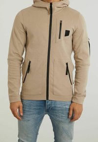 Felpa beige con zip e tasche nere sulla parte anteriore, sul petto e sulla manica, indossata con jeans chiari strappati.