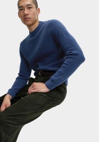 Modrý pletený svetr s vysokým výstřihem, v kombinaci s tmavě zelenými kordovými kalhotami. Svetr má žebrovanou strukturu a přiléhavé manžety.