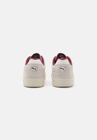 Zapatillas con un upper de ante gris claro y acentos en burdeos; presenta una forma redondeada y un detalle de logo texturizado en el talón.