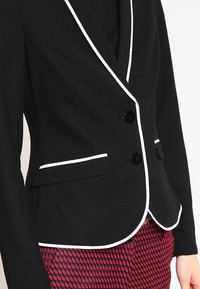 Svart blazer med vit piping, med en figursydd design, två knappar och sidfickor, tillverkad i texturerad tyg.