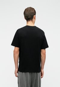 Bărbat îmbrăcat cu un tricou negru simplu cu mânecă scurtă și pantaloni gri, stând cu spatele îndreptat către un fundal alb simplu.