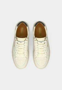 Αθλητικά παπούτσια από κρεμ δέρμα με διάτρητες λεπτομέρειες, επίπεδα κορδόνια και καφέ εσωτερικά πέλματα, με μαύρη και χρυσή σήμανση στη γλώσσα.