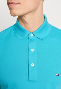 Turkos polo t-shirt med en texturerad tyg, som har en spetsig krage och en knappslå med tre vita knappar. Litet logotypdetalj på bröstet.