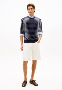 Μπλε ναυτικό και λευκό ριγέ πουλόβερ με στρογγυλή λαιμόκοψη, συνδυασμένο με ανοιχτό μπεζ σορτς και καφέ δερμάτινα παπούτσια με κορδόνια· κομψό, casual καλοκαιρινό ντύσιμο.