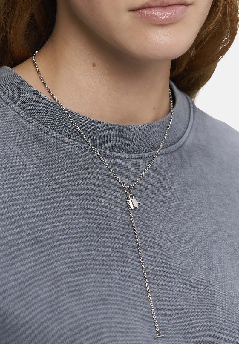 Sølvkæde med et minimalistisk design, der har en lille vedhæng og et stilrent vertikalt drop. Båret over en grå sweatshirt.