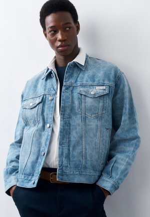 Ung mand iført en lyseblå Calvin Klein denimjakke over en hvid skjorte, stående mod en ensartet lys baggrund med hænderne i lommerne.