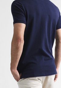 Man wearing a navy blue short-sleeve polo shirt and light beige pants, shown from the back with hands in pockets.