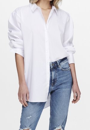 Woman wearing oversized white button-up shirt with rolled sleeves and ripped blue jeans, standing against white background.