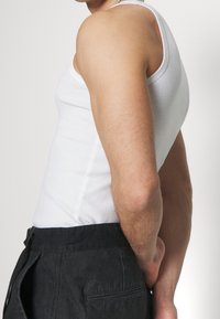 Vit ärmlös linne visat på en person, ihop med mörka byxor. Tyget ser slätt ut, den figurnära designen framhäver formen.