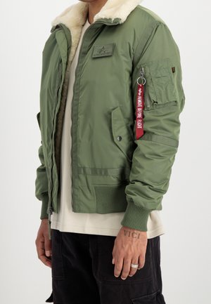 Person trägt grüne Bomberjacke mit weißem Lammfellkragen, rotem "Remove Before Flight" Anhänger an der Ärmeltasche und schwarze Hose.