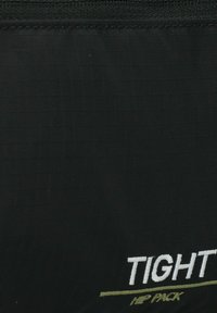 Tejido negro con texto bordado en blanco "TIGHT" y un texto más pequeño en dorado "HIP PACK" debajo, mostrando detalles de textura y costura.