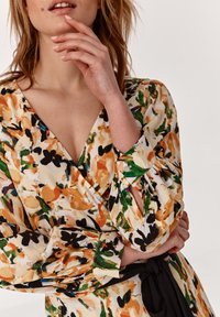 Kvetinové šaty vo farbe smotany s oranžovými, zelenými a čiernymi vzormi. Dlhé puff rukávy s bodkovými akcentmi a detailom viazania v páse.
