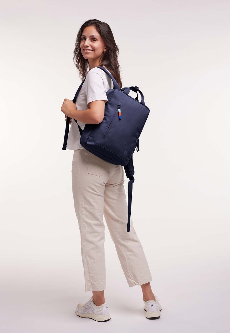 Jeune femme souriante, portant un t-shirt blanc, un pantalon beige, des baskets blanches, avec un sac à dos bleu marine sur le dos, devant un fond uni.