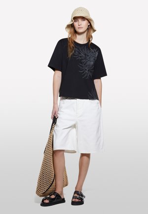 T-shirt nera a maniche corte con design floreale, pantaloni corti bianchi fino al ginocchio, sandali neri e cappello a secchiello lavorato all'uncinetto beige. Borsa intrecciata in mano.