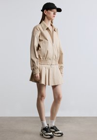 Μπεζ ζακέτα με φερμουάρ, ελαστική μέση και φουσκωτά μανίκια, συνδυασμένη με πλισέ φούστα. Μαύρο καπέλο, πλεκτές κάλτσες και αθλητικά παπούτσια ολοκληρώνουν την εμφάνιση.