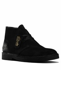 Black suede desert boot with rounded toe, lace-up front, contrasting smooth heel, and stitched detailing. Brand tag on the side.