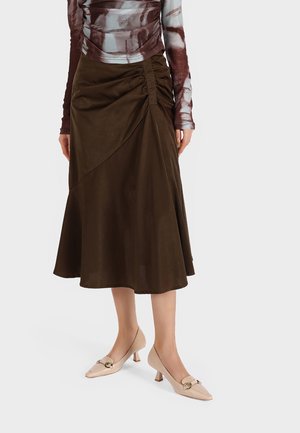 Gonna marrone fino al ginocchio con un design drappeggiato e arricciature laterali, abbinata a tacchi beige con tacco piccolo e dettagli decorativi.
