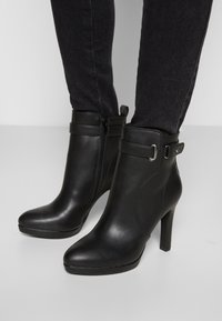 Black leather ankle boots with a glossy finish, featuring a high heel, zip closure, and strap detail near the ankle.