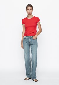 Rotes, tailliertes T-Shirt mit grauen Abschlüssen, kombiniert mit hellblauen, geraden Jeans, getragen mit schwarzen Sandalen mit weißen Akzenten.