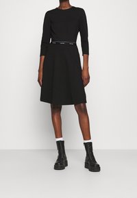 Černé šaty s dlouhým rukávem a rozšířenou sukní, s páskem s logem. Nalezené na černých šněrovacích botách a bílých ponožkách. Hladký povrch.