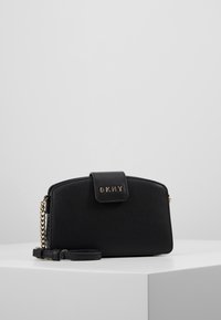 Wristlet DKNY in pelle nera testurizzata con dettagli in catena dorata e chiusura a pattina, esposto su un piedistallo bianco contro uno sfondo grigio chiaro.