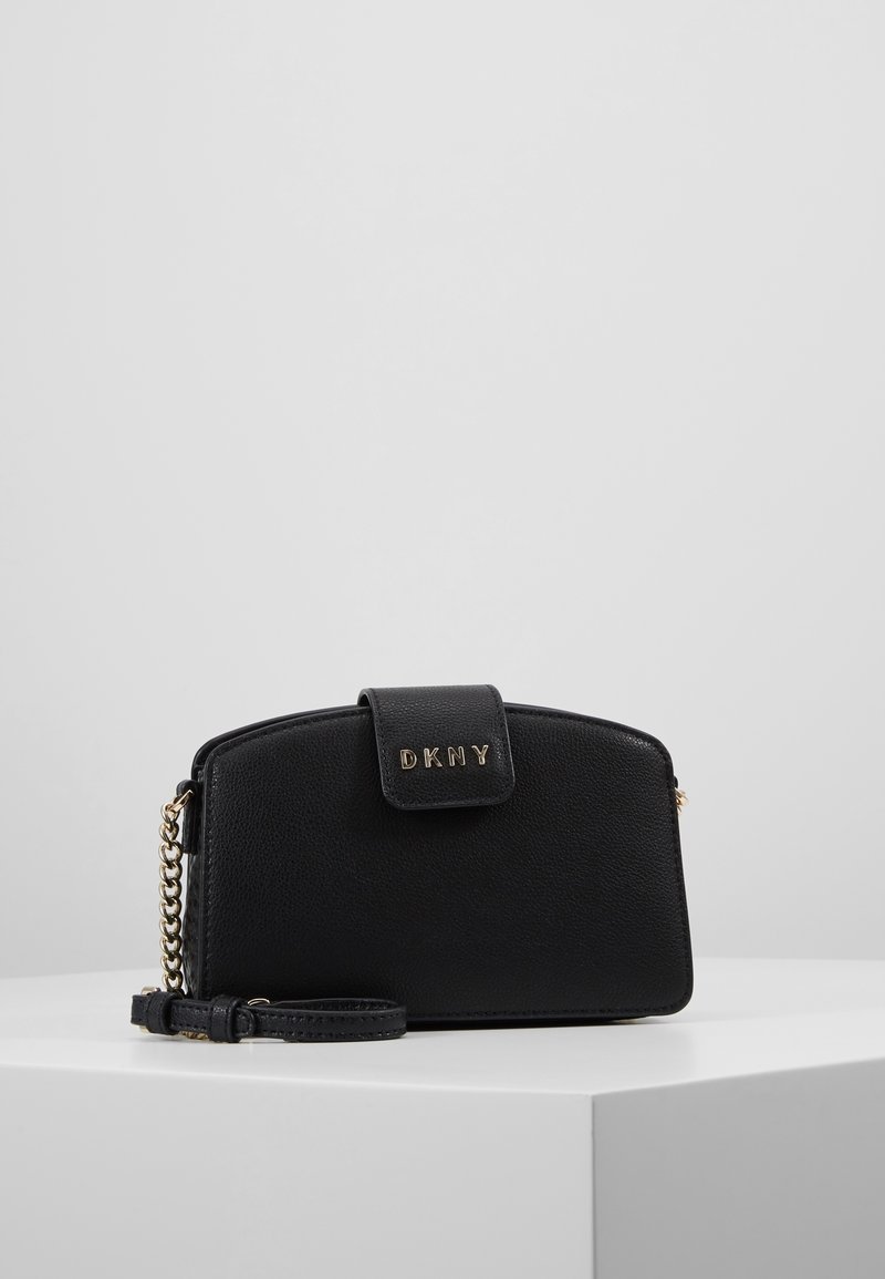 Wristlet DKNY in pelle nera testurizzata con dettagli in catena dorata e chiusura a pattina, esposto su un piedistallo bianco contro uno sfondo grigio chiaro.